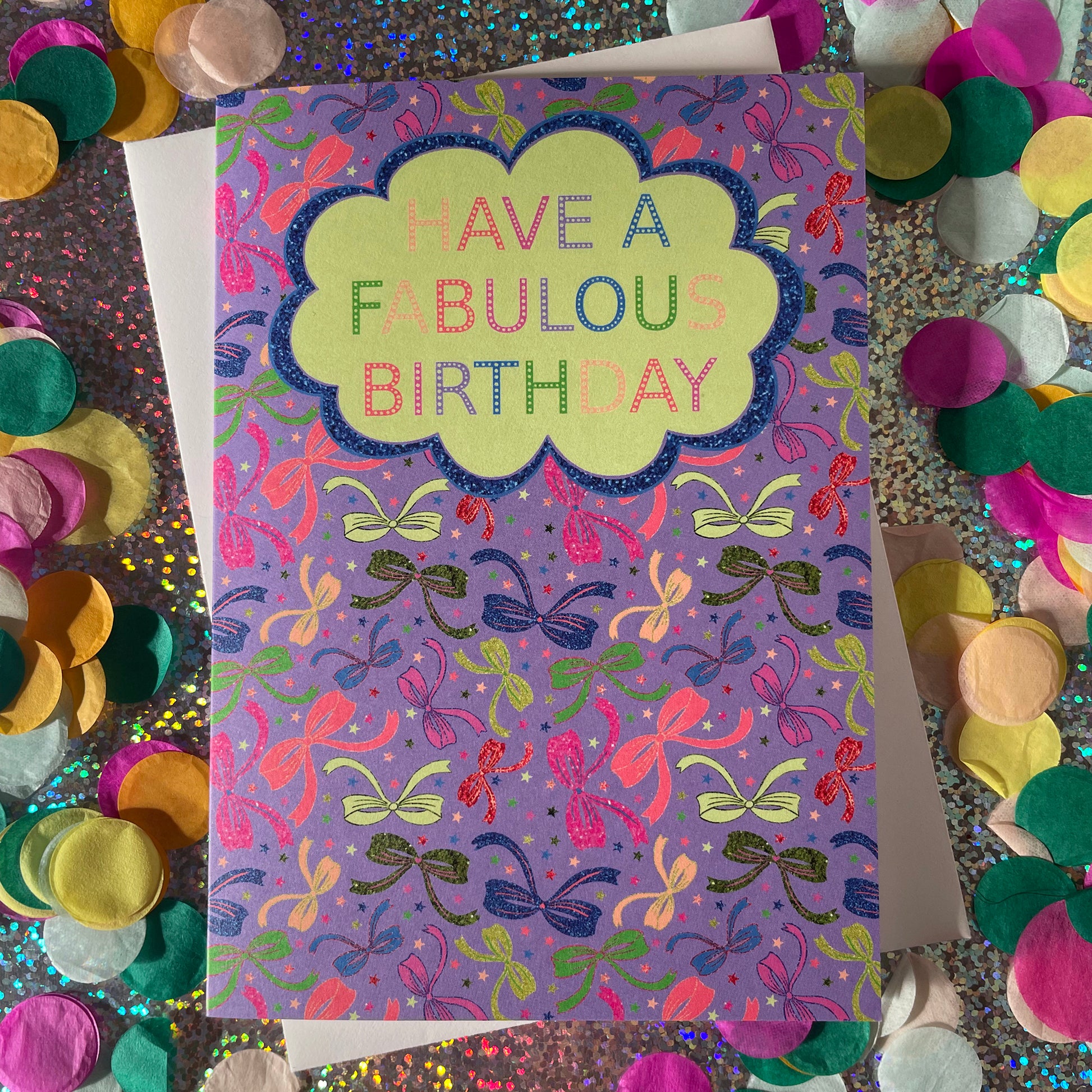 Image shows a lilac birthday card with neon and sparkly bow illustrations and a cloud with multicoloured wording that reads 'Have a fabulous birthday"
