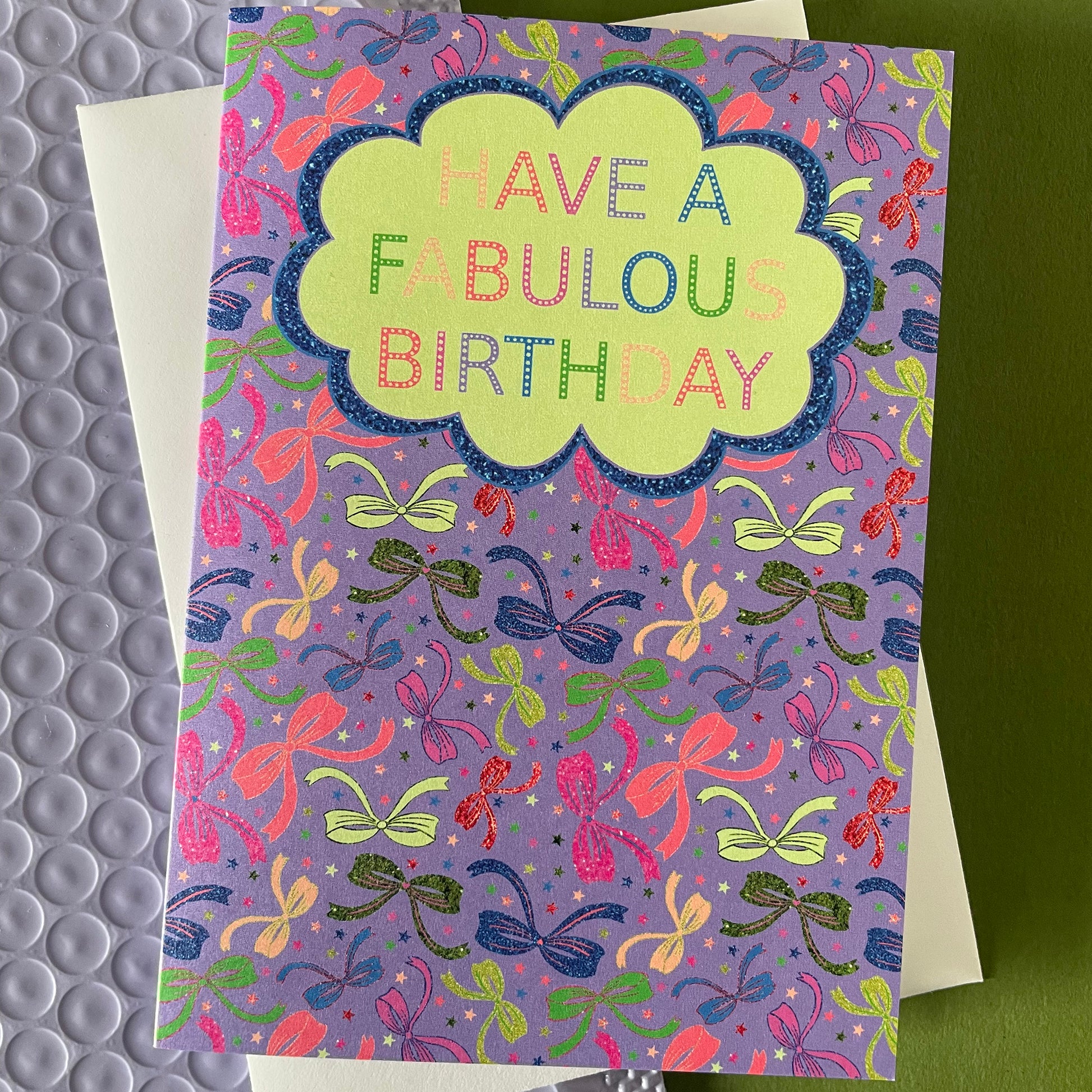 Image shows a lilac birthday card with neon and sparkly bow illustrations and a cloud with multicoloured wording that reads 'Have a fabulous birthday"