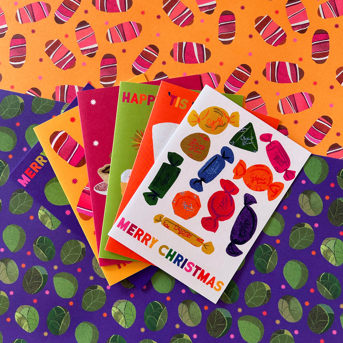 A stack of colourful Christmas greeting cards fanned out on top of patterned wrapping paper. The front card is white with illustrations of wrapped chocs and the text 'MERRY CHRISTMAS' in rainbow letters.