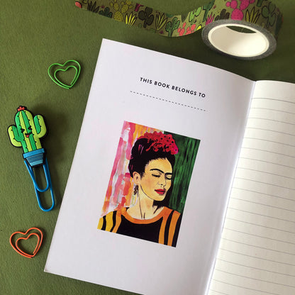 Image shows an open notebook featuring a bright, hand painted illustration of Frida Kahlo on the inside cover and lined pages