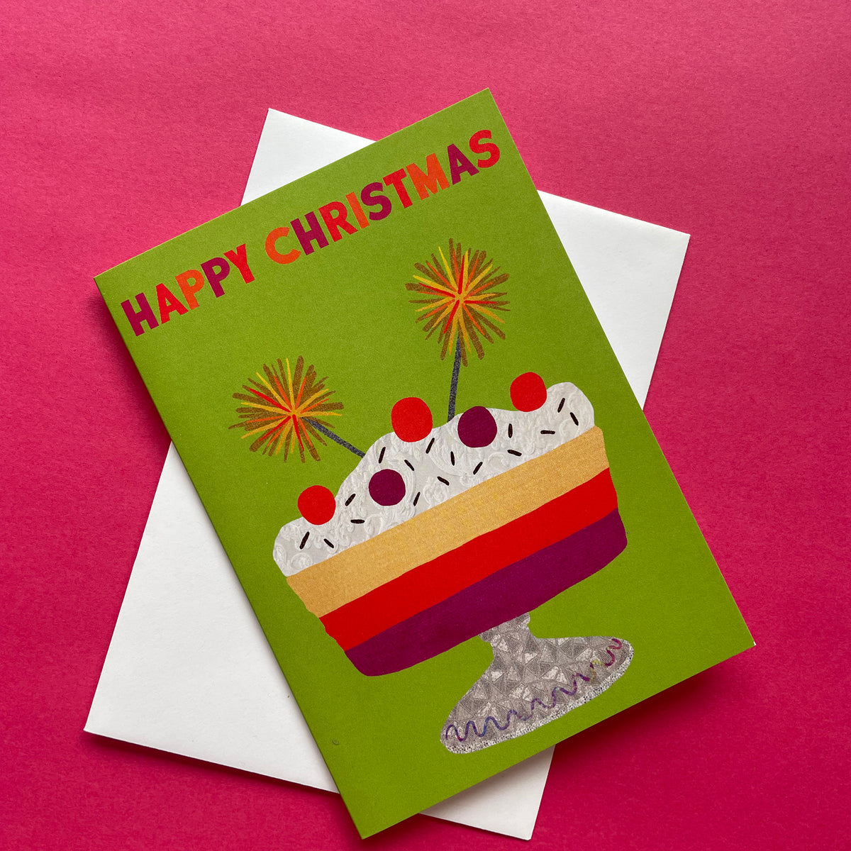 A Christmas greeting card with a green background and a colourful dessert illustration, placed on top of a white envelope against a bright pink background.