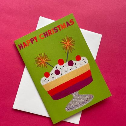 A Christmas greeting card with a green background and a colourful dessert illustration, placed on top of a white envelope against a bright pink background.