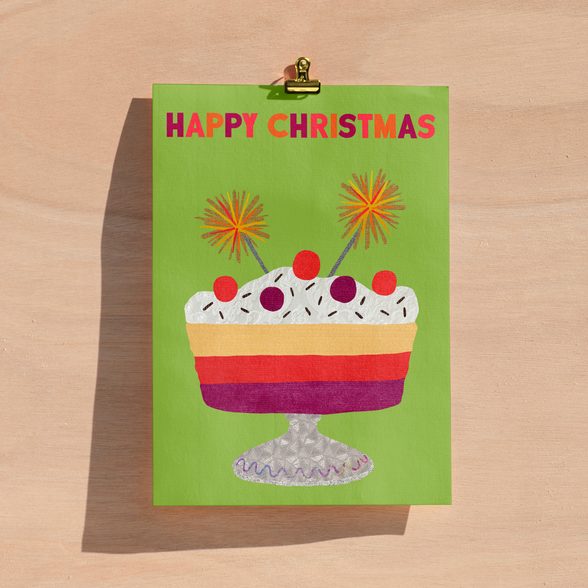 A green Christmas greeting card with a colourful layered dessert illustration and the words 'HAPPY CHRISTMAS' in pink and orange, clipped at the top with a gold metal clip against a light wood background.
