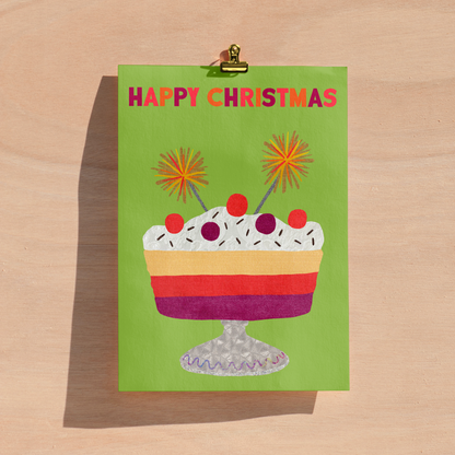 A green Christmas greeting card with a colourful layered dessert illustration and the words 'HAPPY CHRISTMAS' in pink and orange, clipped at the top with a gold metal clip against a light wood background.