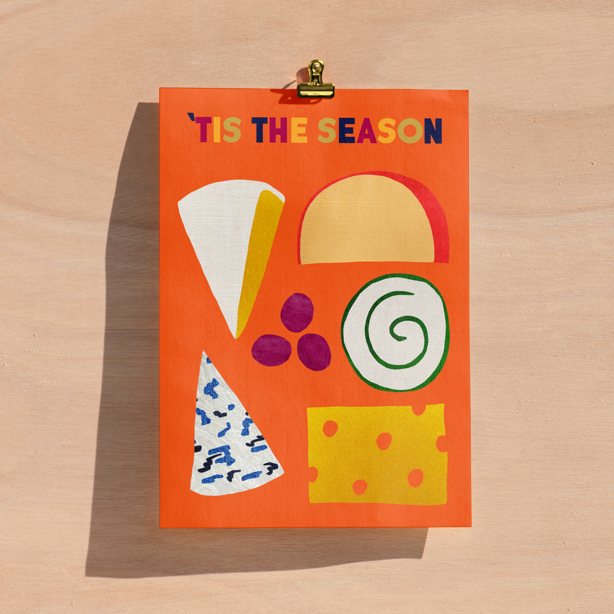 An orange art print with the text "'TIS THE SEASON" in multicoloured letters, decorated with hand-drawn illustrations of cheese wedges, a yellow cheese block, purple grapes, and a garlic cheese spiral, clipped to a light wood background.