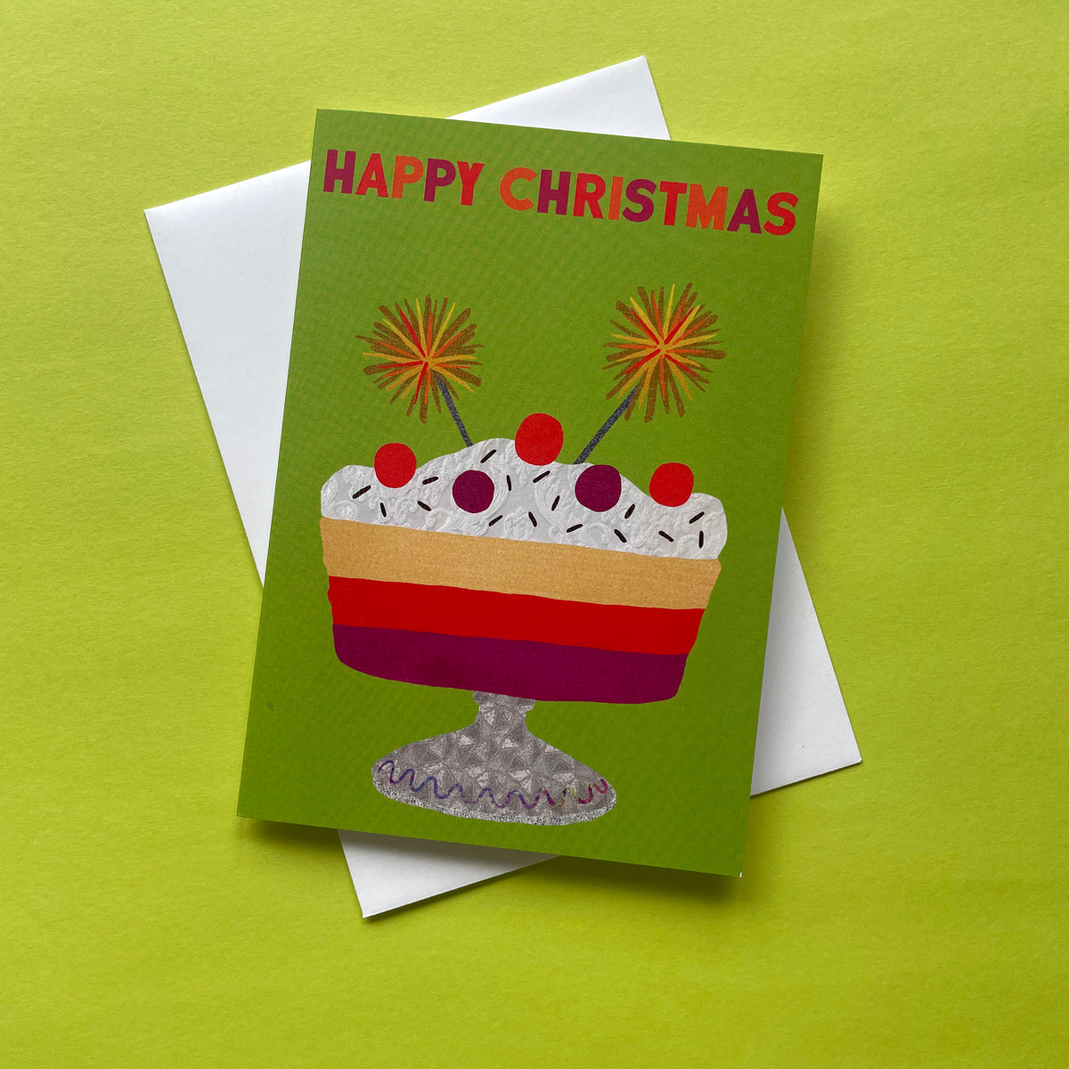 A Christmas greeting card with a green background and a colourful dessert illustration, shown with a white envelope on a bright yellow-green surface. The card reads 'HAPPY CHRISTMAS' in red letters above the dessert.