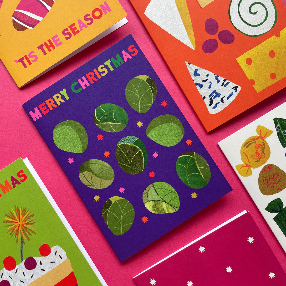 A styled flat-lay of Christmas greeting cards arranged on a bright pink background. The central card is dark purple with green sprout shapes and the words 'MERRY CHRISTMAS' in pink, white, and green.