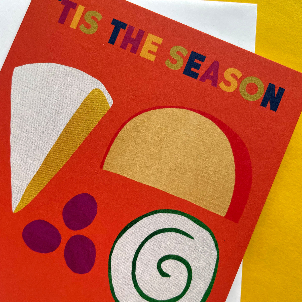 A close-up of a colourful Christmas greeting card with the text 'TIS THE SEASON' in multicoloured letters. The card features cheese and grapes in red, gold, white, purple, and green on a bright red-orange background, layered over a yellow and white textured surface.