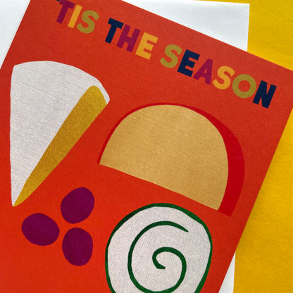 A close-up of a colourful Christmas greeting card with the text 'TIS THE SEASON' in multicoloured letters. The card features cheese and grapes in red, gold, white, purple, and green on a bright red-orange background, layered over a yellow and white textured surface.