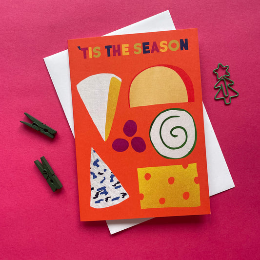 A bright orange greeting card with colourful cheese illustrations, layered over a white envelope on a pink surface. The card reads “TIS THE SEASON” in multicoloured letters.