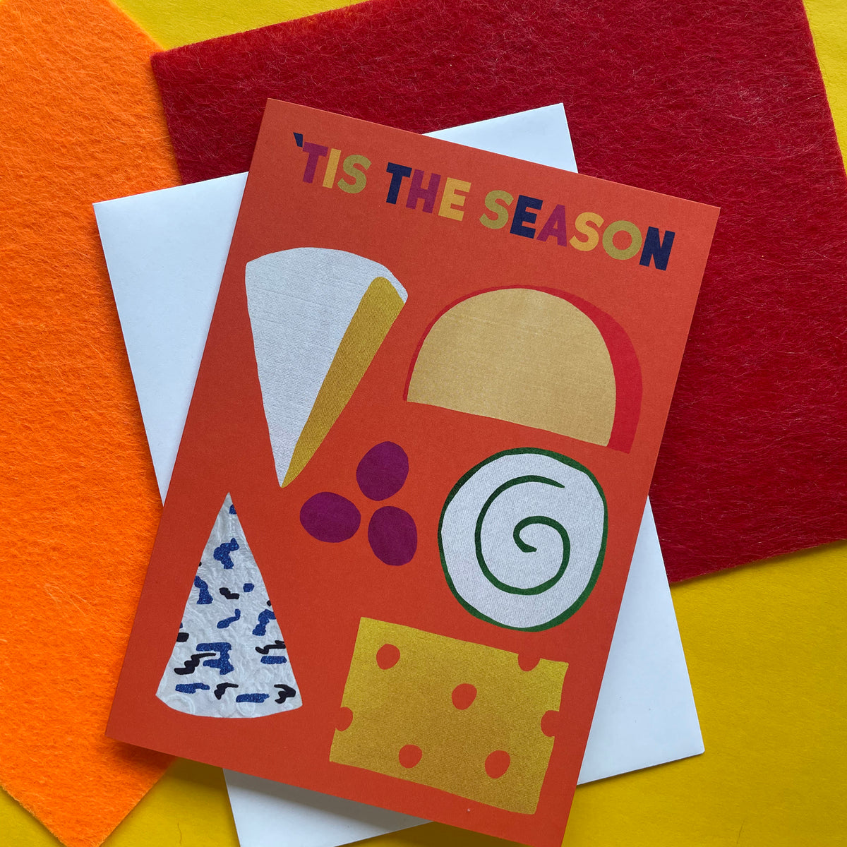 A festive greeting card with the text "'TIS THE SEASON" in multicoloured letters, featuring abstract illustrations of cheese wedges and grapes on an orange-red background. The card is layered over white and red envelopes on a bright yellow surface with orange and red felt paper accents.