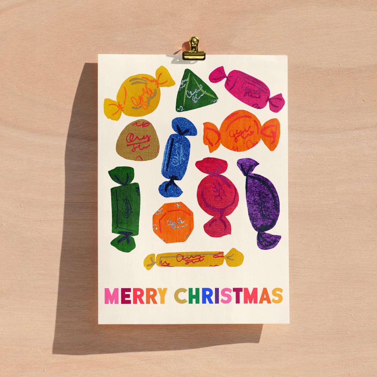 A Christmas greeting card featuring colorful chocolate illustrations and the text 'MERRY CHRISTMAS' in rainbow letters, held by a gold clip on a light wood background.