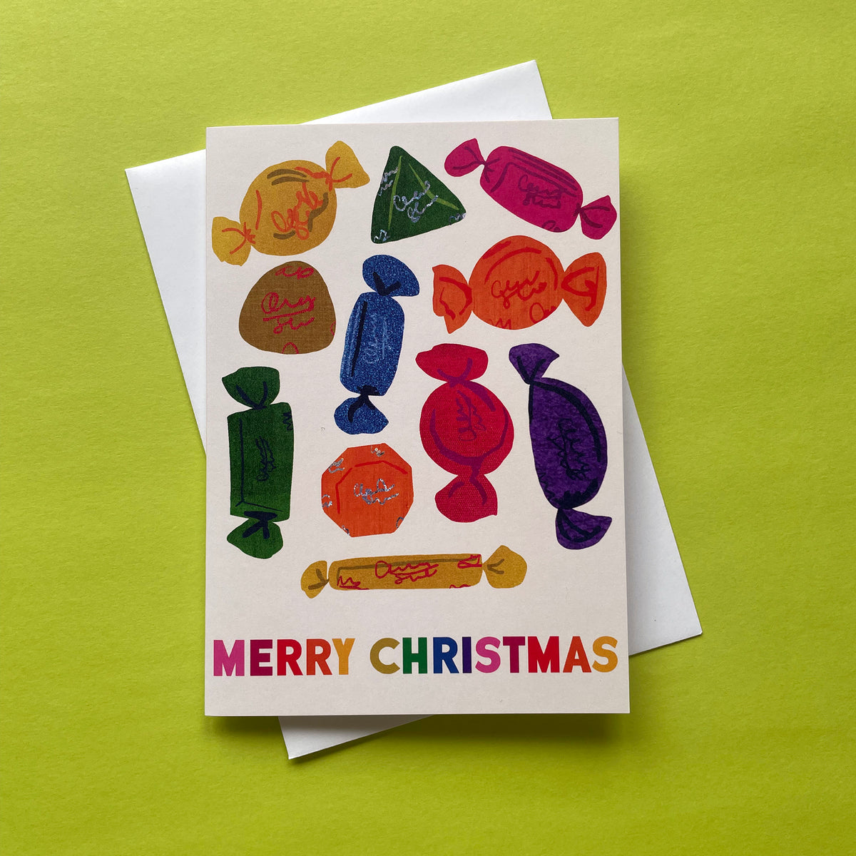 A Christmas greeting card featuring colourful illustrations of wrapped chocolates and the text 'MERRY CHRISTMAS' in rainbow letters, shown with a white envelope on a bright lime green background.