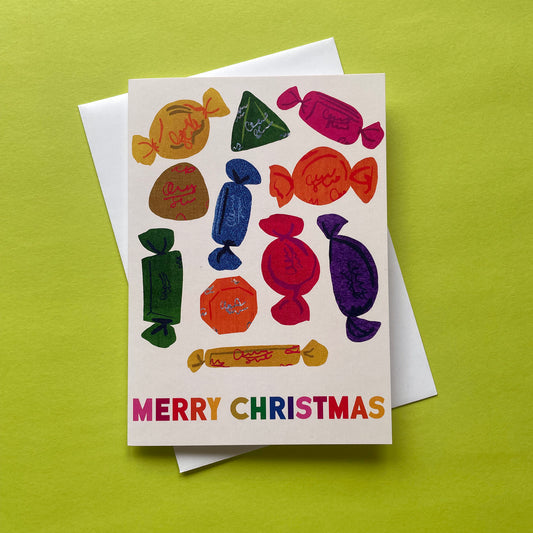 A Christmas greeting card featuring colourful illustrations of wrapped chocolates and the text 'MERRY CHRISTMAS' in rainbow letters, shown with a white envelope on a bright lime green background.