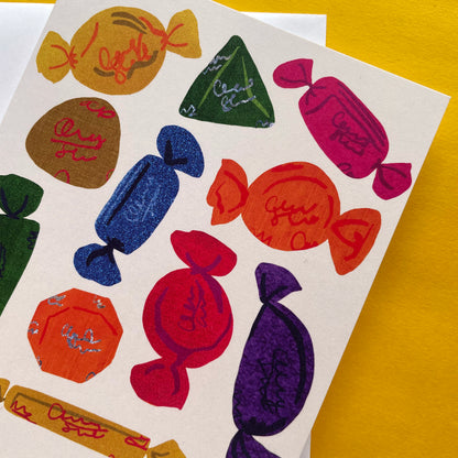 A close-up of a printed card featuring colourful chocolate illustrations in red, orange, blue, green, purple, and pink on a white background, placed on a bright yellow surface.