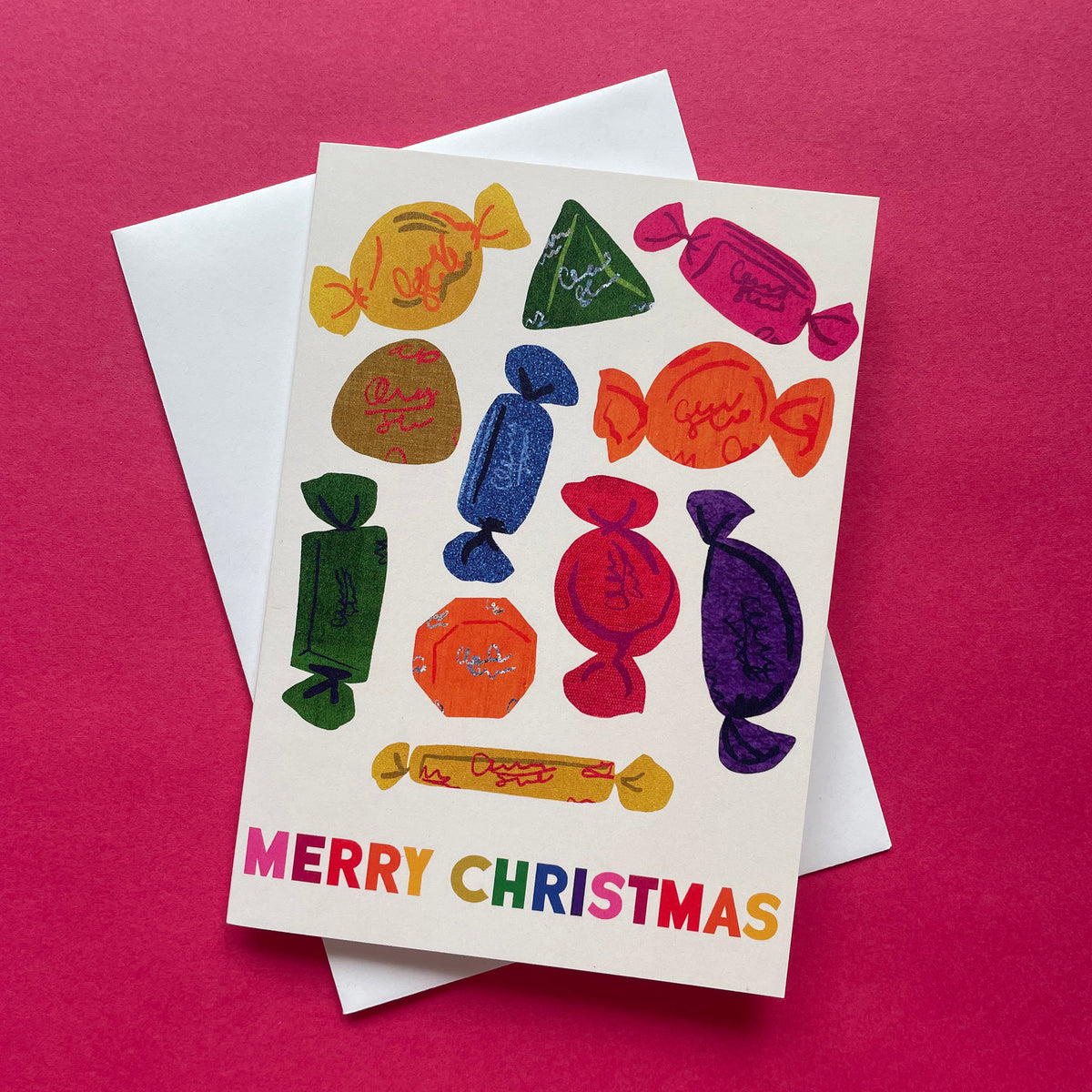 A Christmas greeting card featuring colorful illustrated wrapped chocolates and the text 'MERRY CHRISTMAS' in rainbow letters, shown with a white envelope on a bright pink background.