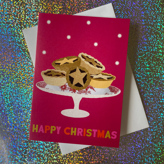 A Christmas greeting card featuring a festive illustration of mince pies on a cake stand against a deep magenta background with white stars. The card reads 'HAPPY CHRISTMAS' in colourful letters and is shown with a white envelope on a glittery multicolour background.
