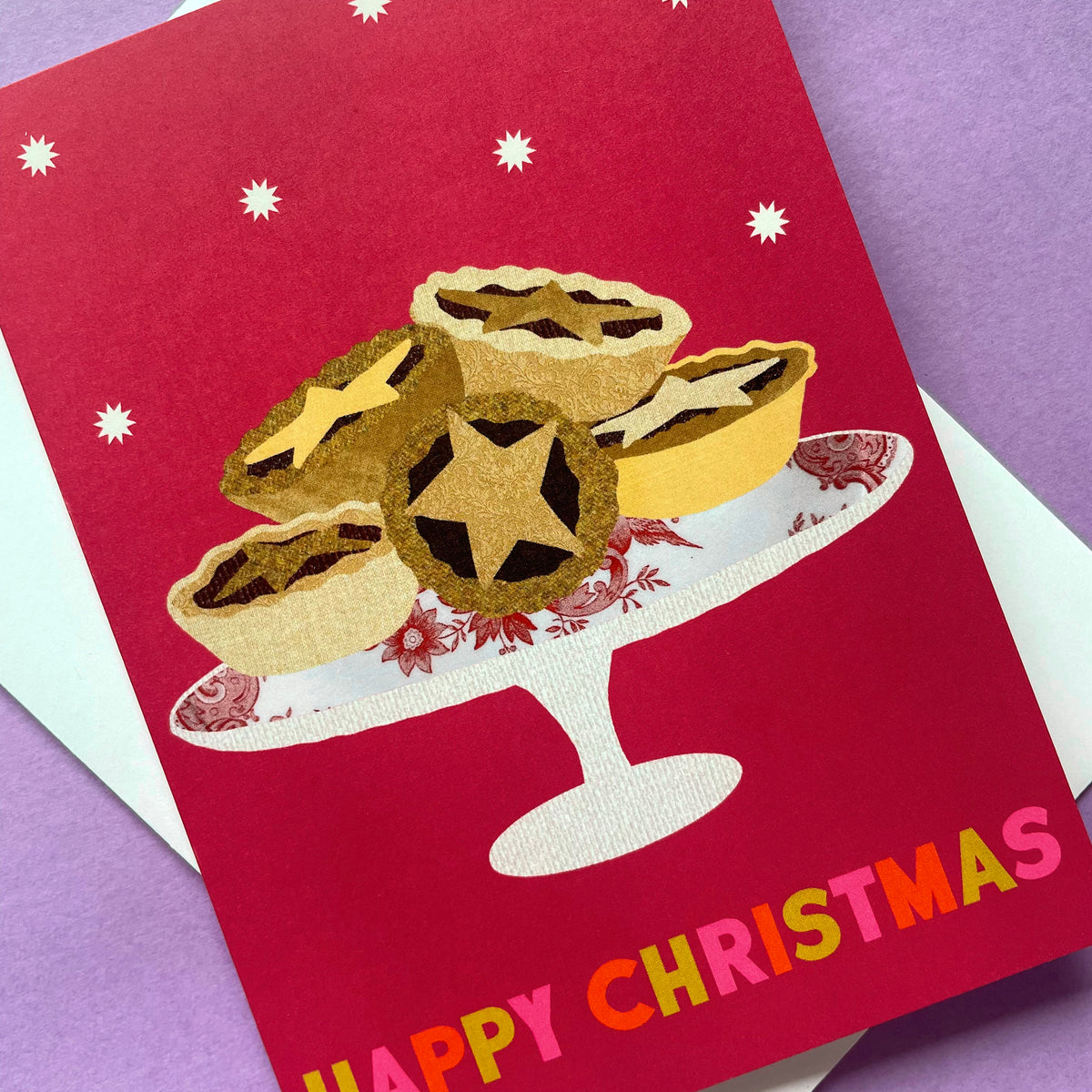 A festive Christmas greeting card featuring an illustration of mince pies on a white pedestal plate against a deep red background, with the words 'HAPPY CHRISTMAS' in colourful letters at the bottom. The card is layered over a white envelope on a purple surface.