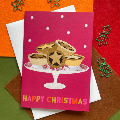 A Christmas greeting card featuring an illustration of mince pies on a stand, with the text 'HAPPY CHRISTMAS' in colourful letters. The card is layered over white and brown envelopes on a festive multicolour felt background with green metal tree cutouts.