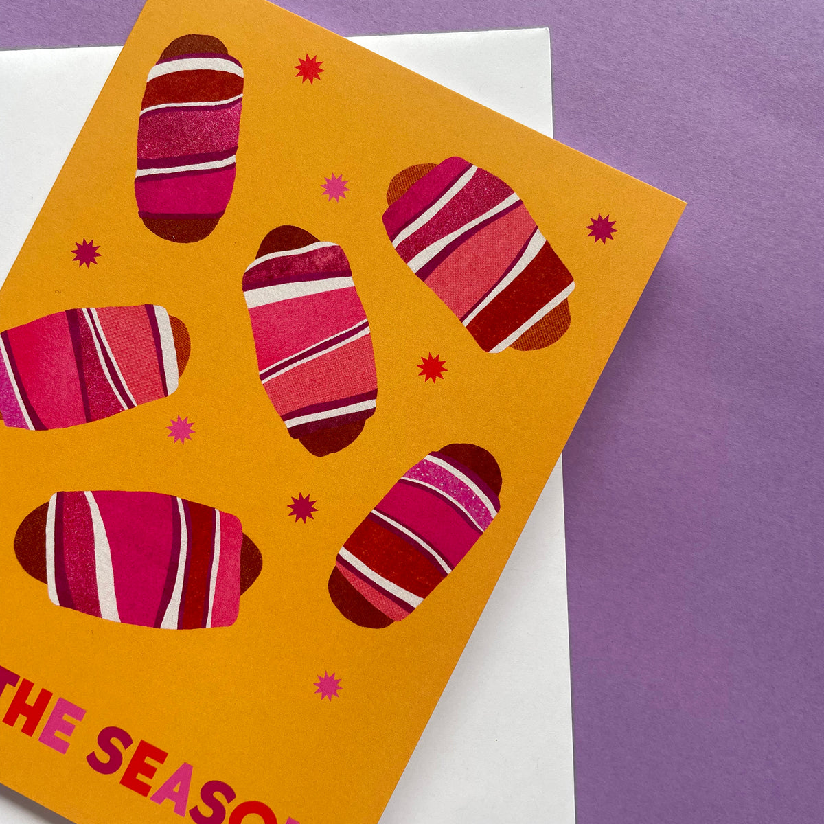 A bright greeting card with a mustard yellow background and stylised pink, and white striped pigs in blanket illustrations, photographed on top of a white card against a purple surface.