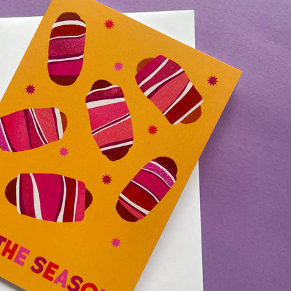 A bright greeting card with a mustard yellow background and stylised pink, and white striped pigs in blanket illustrations, photographed on top of a white card against a purple surface.