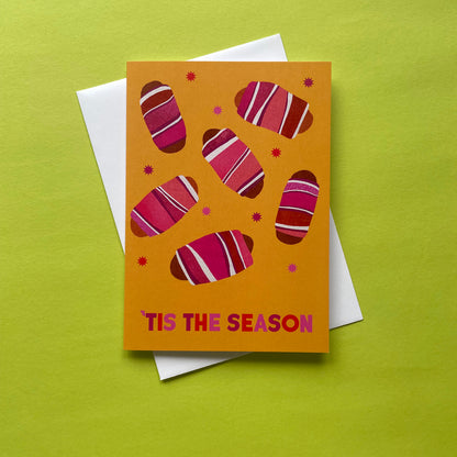 A festive greeting card with a mustard yellow background, decorated with pink pigs in blankets and the text 'TIS THE SEASON' at the bottom. The card is displayed on top of a white envelope against a bright lime green background.