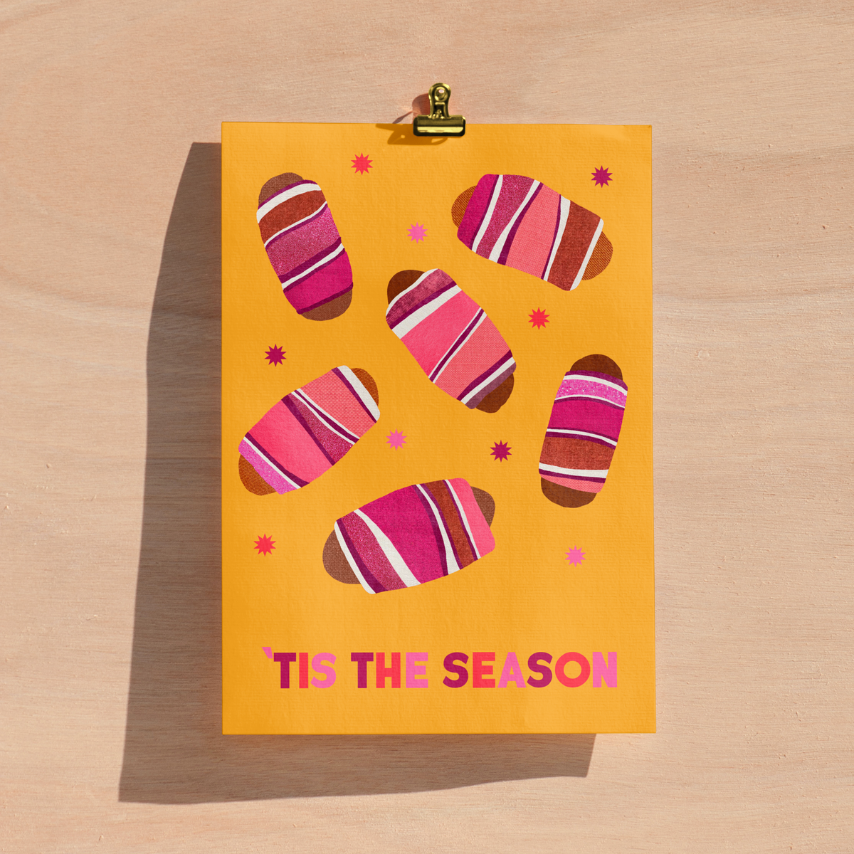A yellow greeting card featuring illustrations of pigs in blankets and the text 'TIS THE SEASON' in pink, clipped at the top with a gold binder clip on a light wood background.