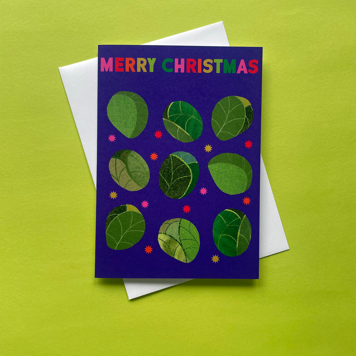 A Christmas greeting card with a dark purple background, featuring the words 'MERRY CHRISTMAS' in multicoloured letters at the top. The card is decorated with green sprout illustrations and small pink and yellow star accents.