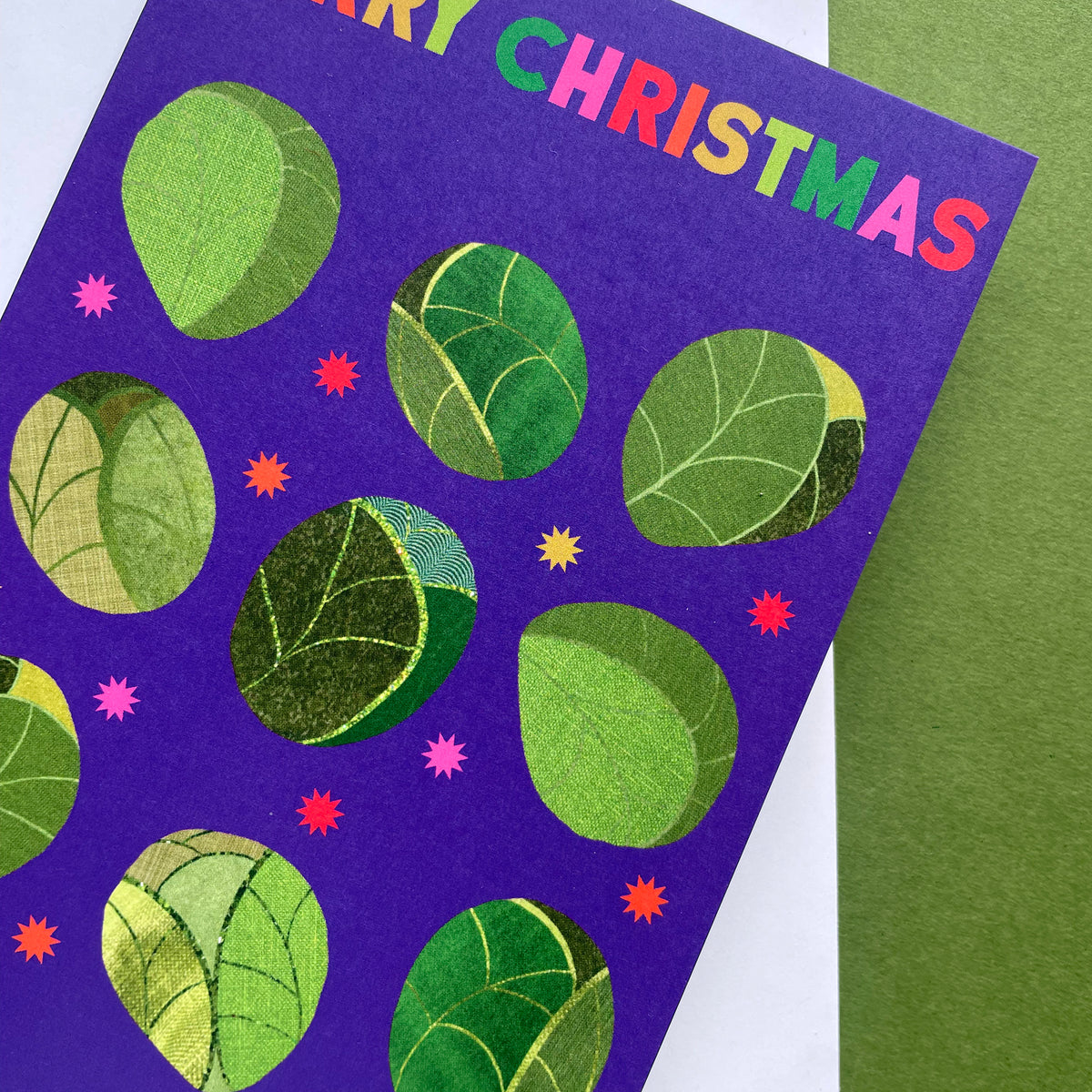 A close-up of a purple Christmas greeting card featuring green sprouts and colourful text reading 'MERRY CHRISTMAS' at the top, with a green textured surface behind it.