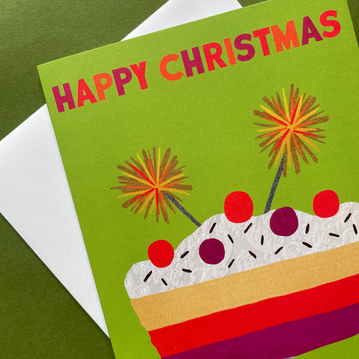 A Christmas greeting card with a bright green background, reading 'HAPPY CHRISTMAS' in pink and orange letters above a stylised layered trifle with red, purple, beige, and white details. The card is photographed at an angle over a white envelope on a dark green surface.