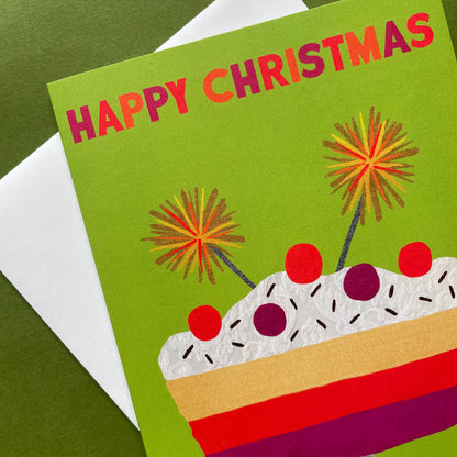 A Christmas greeting card with a bright green background, reading 'HAPPY CHRISTMAS' in pink and orange letters above a stylised layered trifle with red, purple, beige, and white details. The card is photographed at an angle over a white envelope on a dark green surface.