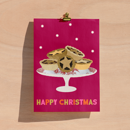 A Christmas greeting card with a bright magenta background, featuring an illustration of mince pies on a white cake stand and the words 'HAPPY CHRISTMAS' in multicoloured letters. The card is held at the top by a gold clip against a light wood background.