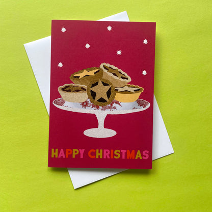 A Christmas greeting card featuring an illustration of mince pies on a stand, with the text 'HAPPY CHRISTMAS' at the bottom, placed on top of a white envelope against a bright lime green background.