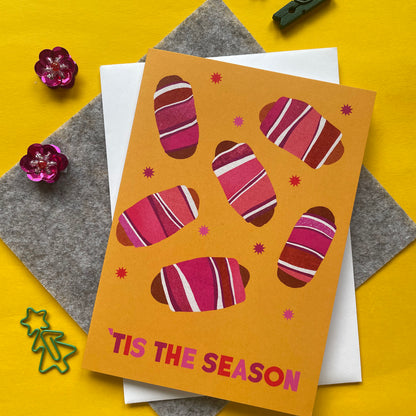 A greeting card with a mustard yellow background and illustrated striped pigs in blankets, layered over a white envelope on grey felt sheets, with small decorative flowers and star cutouts on a bright yellow surface. The card reads “TIS THE SEASON.