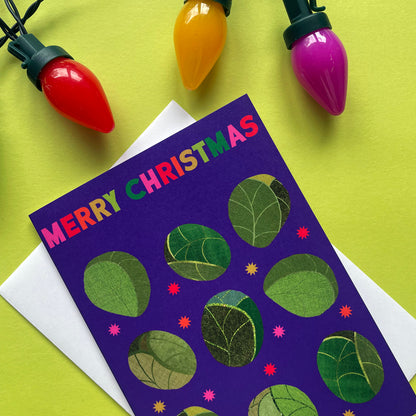A Christmas greeting card with 'MERRY CHRISTMAS' in colourful letters, decorated with sprouts on a purple background, placed on a white envelope over a bright yellow-green surface with hanging multicolour Christmas bulbs.