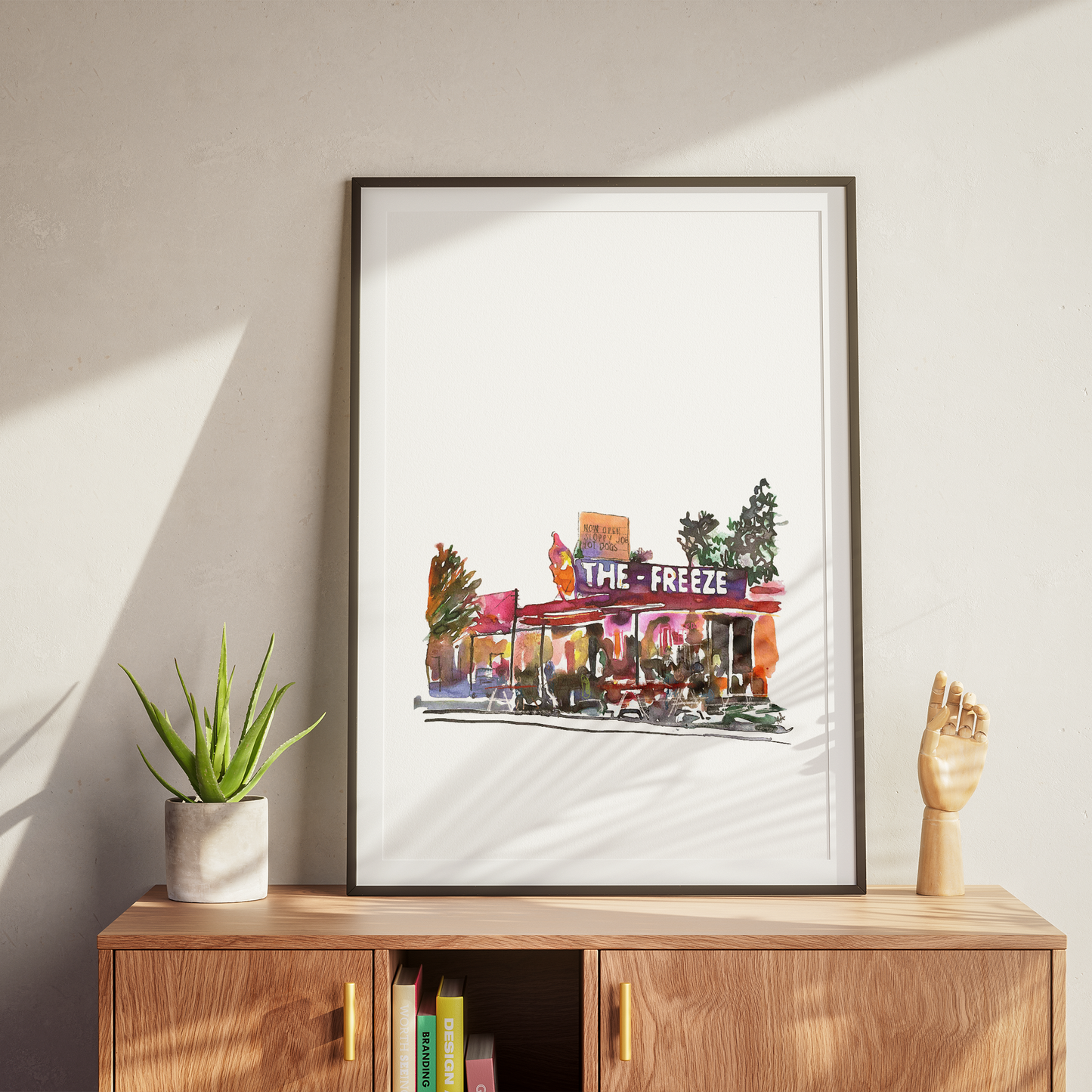 Framed artwork of a city street scene titled 'The Freeze' on a wooden cabinet with a plant and books.
