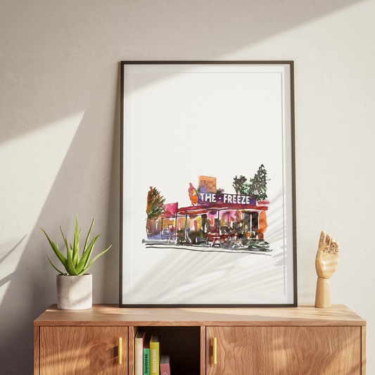 Framed artwork of a city street scene titled 'The Freeze' on a wooden cabinet with a plant and books.