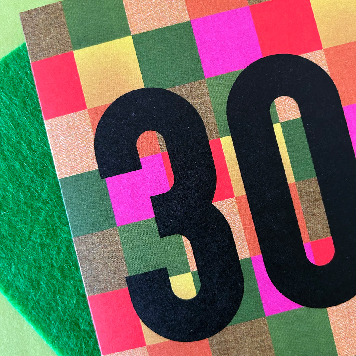 A close-up of a colourful square card or paper featuring a multicolour checkered pattern with large black numbers '30' overlaid, placed on top of a bright green felt surface.