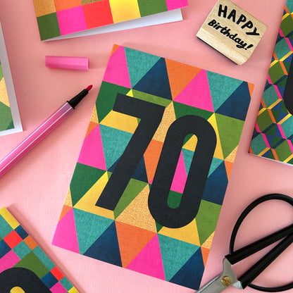 A colourful birthday card with a geometric triangle pattern and the number 70 in large black letters, styled on a pink background with scissors, a highlighter, and other craft items.