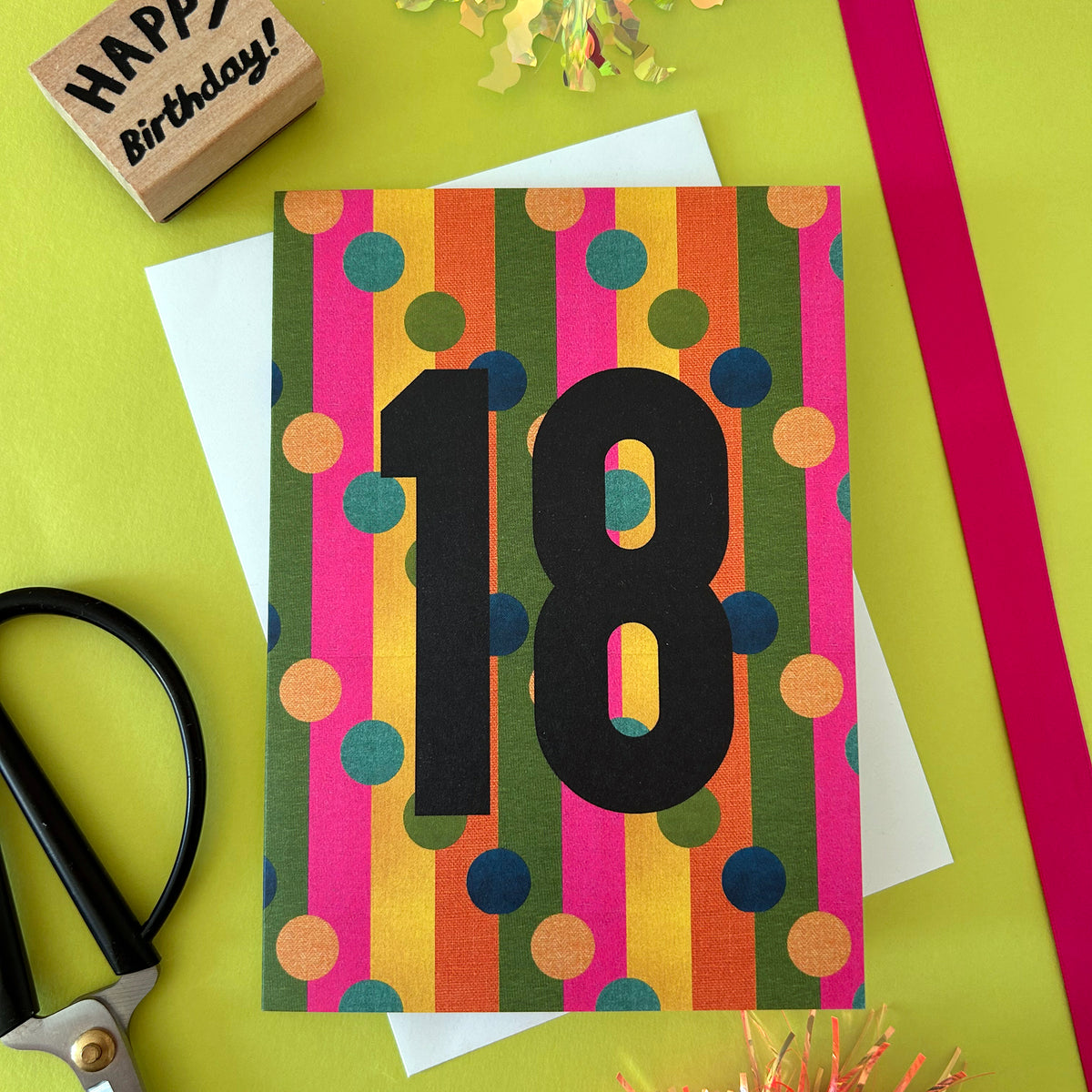 A colourful birthday card with vertical stripes in pink, orange, yellow, green, and blue, overlaid with a large black number 18. The card is styled on a bright yellow-green background with craft props including scissors, red ribbon, a wooden rubber stamp that says 'HAPPY Birthday!', and a white envelope.