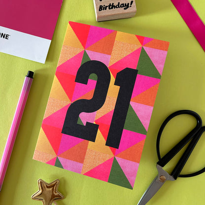 A colourful birthday card with a geometric triangle pattern in pink, orange, yellow, and green, featuring the number '21' in large black font. The card is styled in a flat-lay arrangement on a bright yellow-green background with scissors, a pink pen, a gold star, a rubber stamp, and other stationery items.