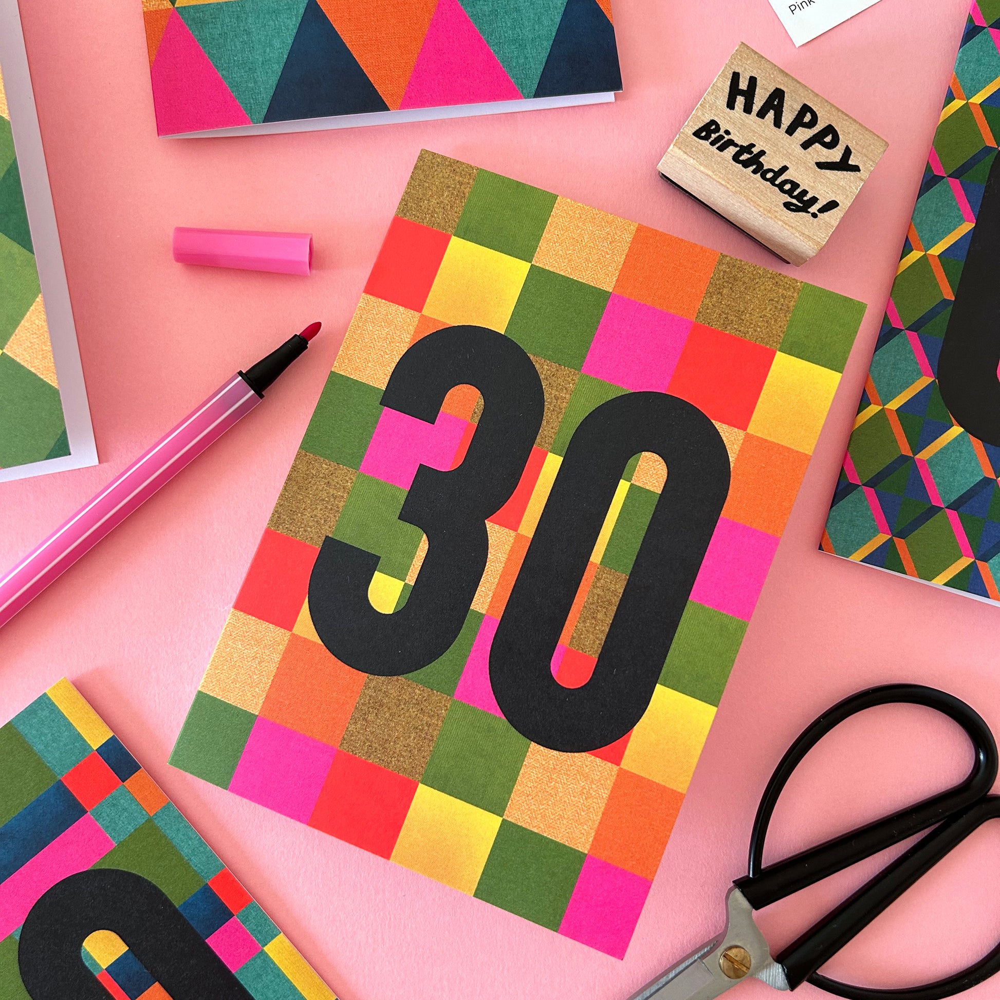A colourful birthday card with a multicolour checkered pattern and large black '30' text, styled on a pink background with scissors, a pink marker, a wooden stamp, and other card designs.