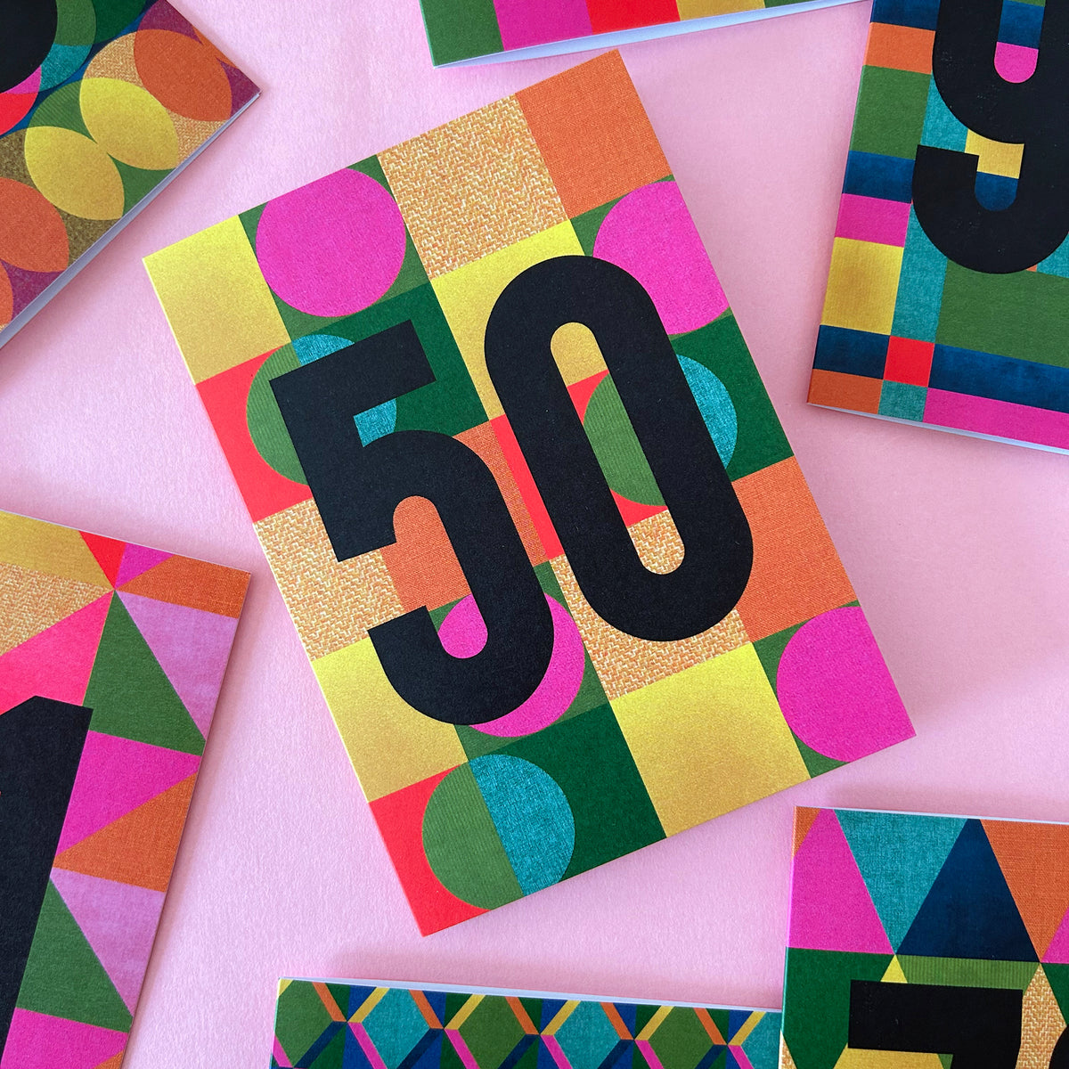A colourful geometric 50th birthday card with bold black '50' text, surrounded by other matching patterned cards on a pink background.