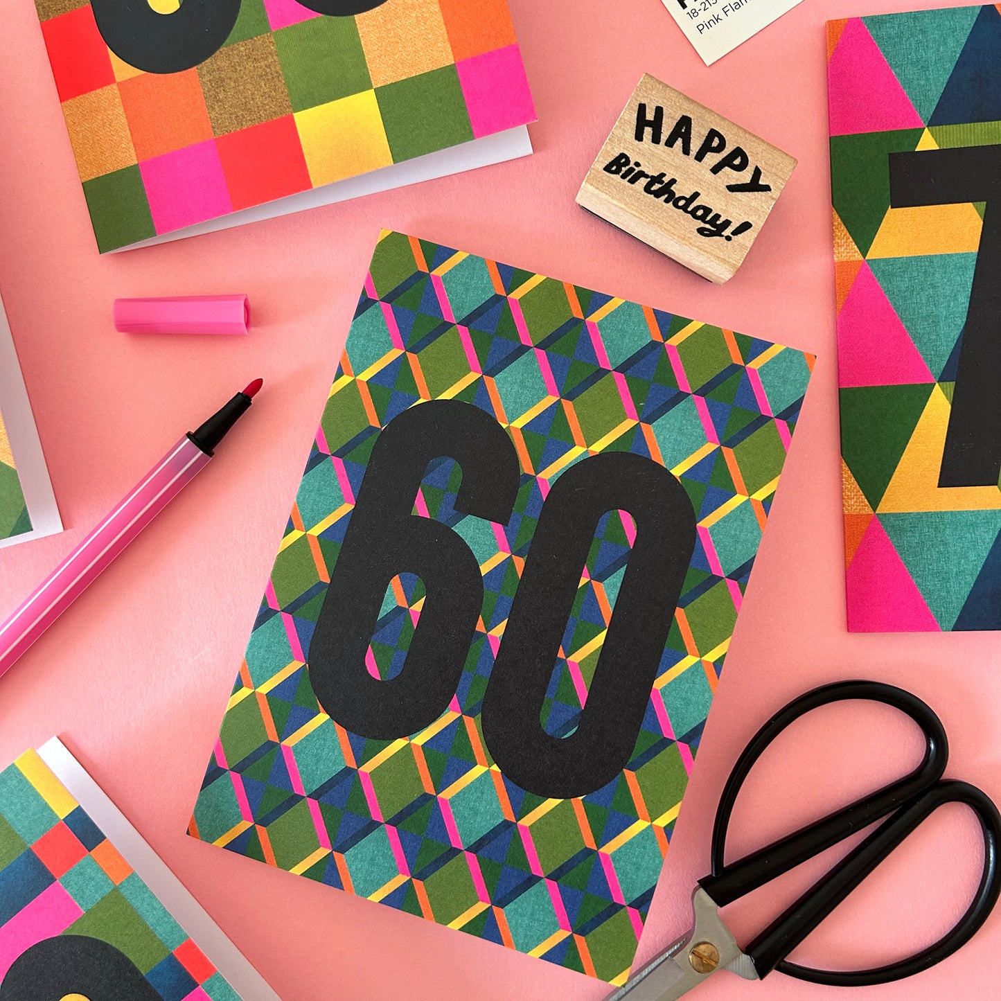 A colourful birthday card with a geometric pattern and large black '60' text, styled on a pink background with scissors, a pink marker, a wooden 'Happy Birthday!' stamp, and other matching cards.