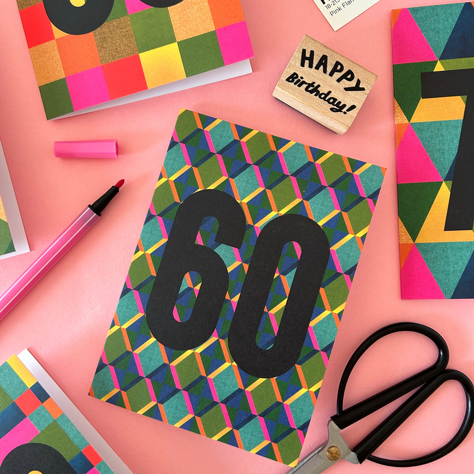 A colourful birthday card with a geometric pattern and large black '60' text, styled on a pink background with scissors, a pink marker, a wooden 'Happy Birthday!' stamp, and other matching cards.