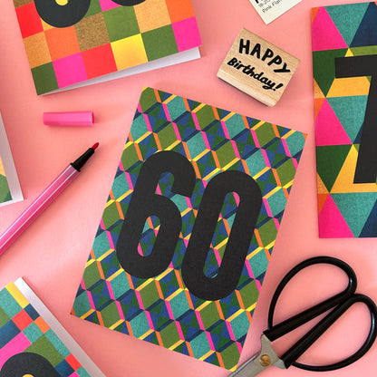 A colourful birthday card with a geometric pattern and large black '60' text, styled on a pink background with scissors, a pink marker, a wooden 'Happy Birthday!' stamp, and other matching cards.