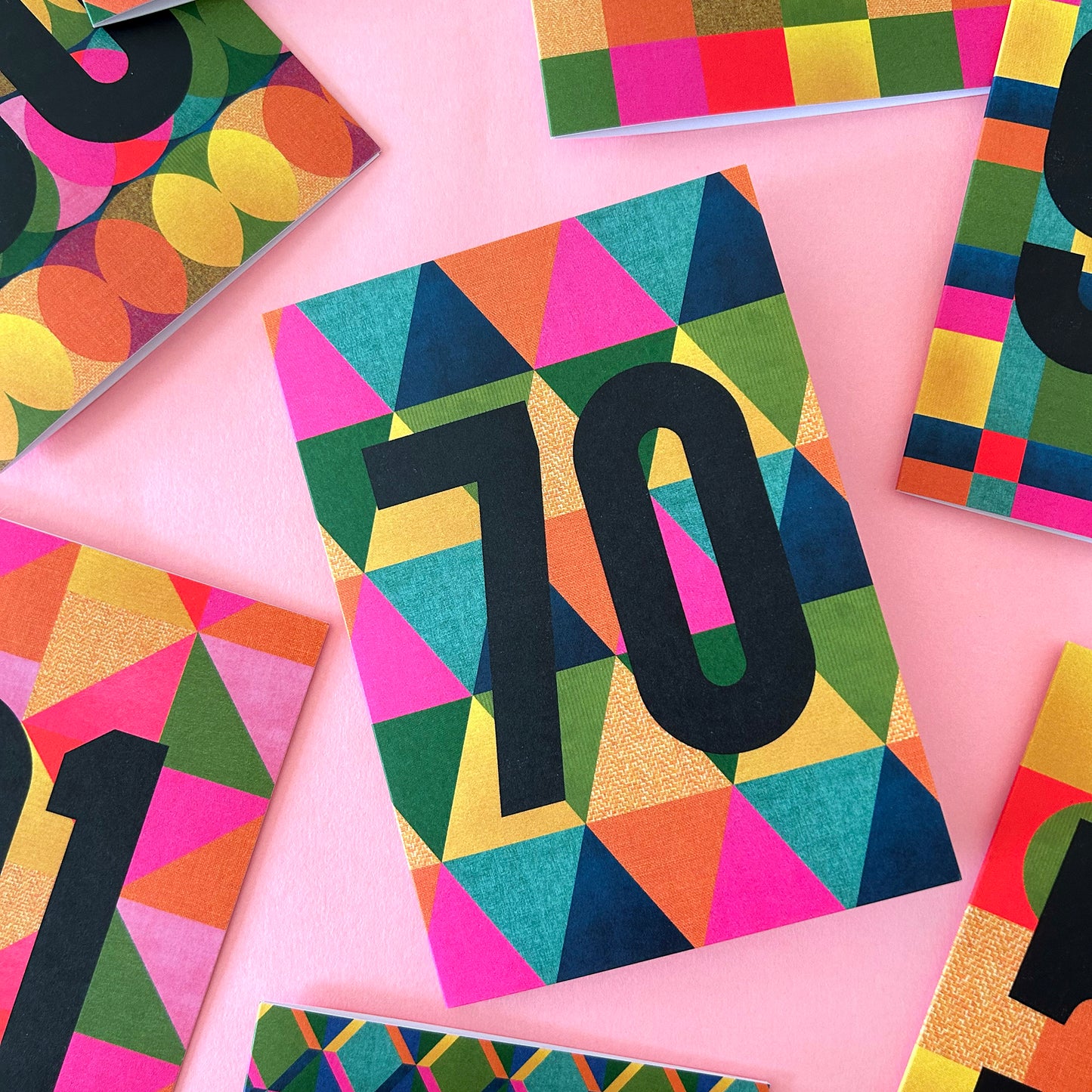 A colourful geometric greetings card featuring the number '70' in bold black font over a vibrant multicolour triangle pattern, styled on a pink background with other cards partially visible.