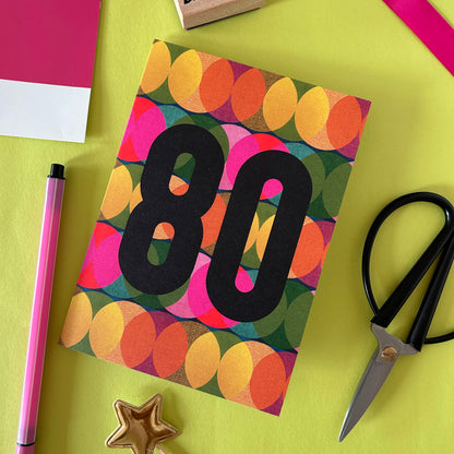 A colourful greeting card with a retro pattern of overlapping circles in orange, yellow, pink, green, and blue, featuring the number '80' in bold black text. The card is styled in a flat-lay arrangement on a bright yellow-green background with a pink pen, scissors, and a gold star.
