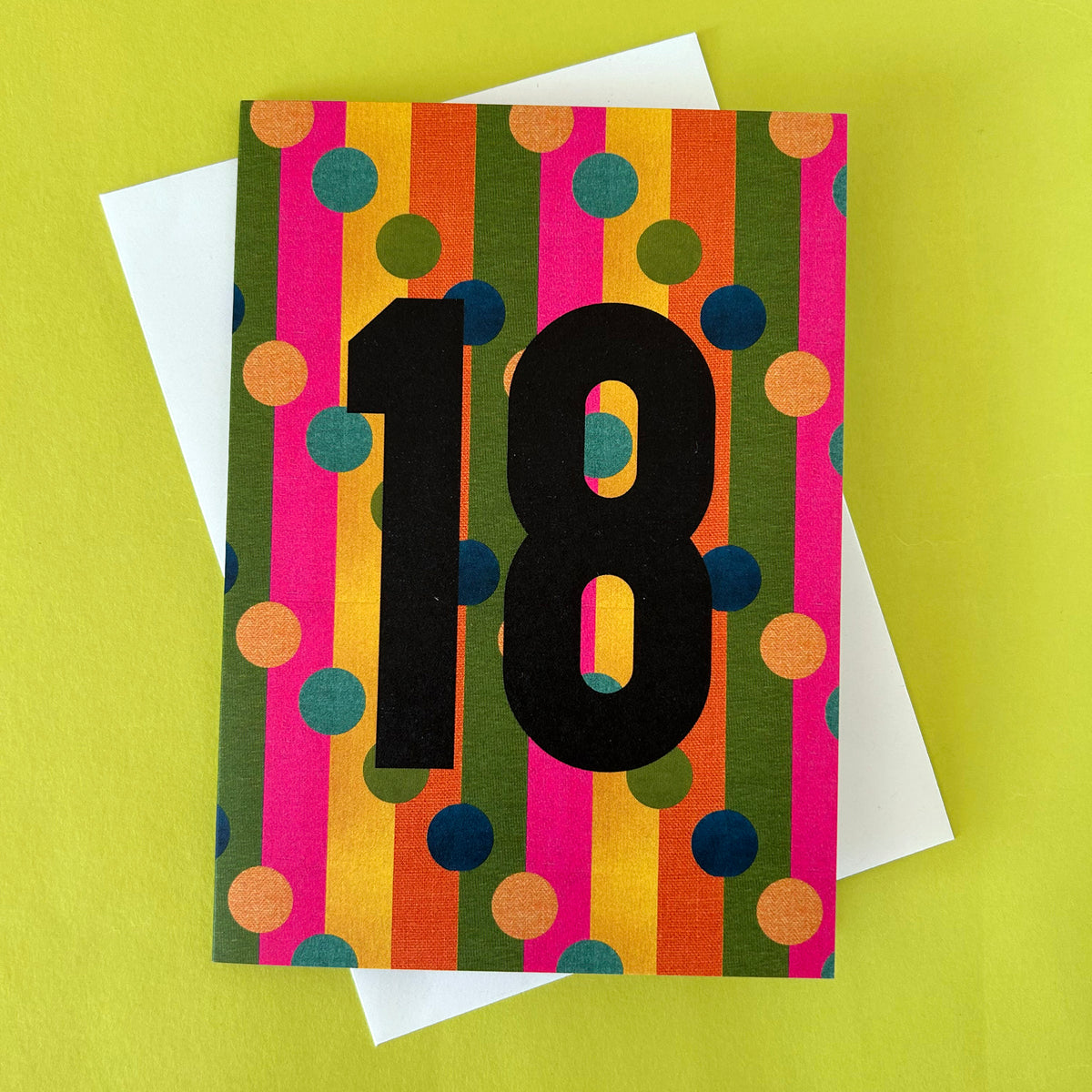 A colourful 18th birthday greeting card with bold black numerals '18' over vertical stripes of pink, orange, yellow, green, and blue polka dots, shown with a white envelope on a bright yellow-green background.