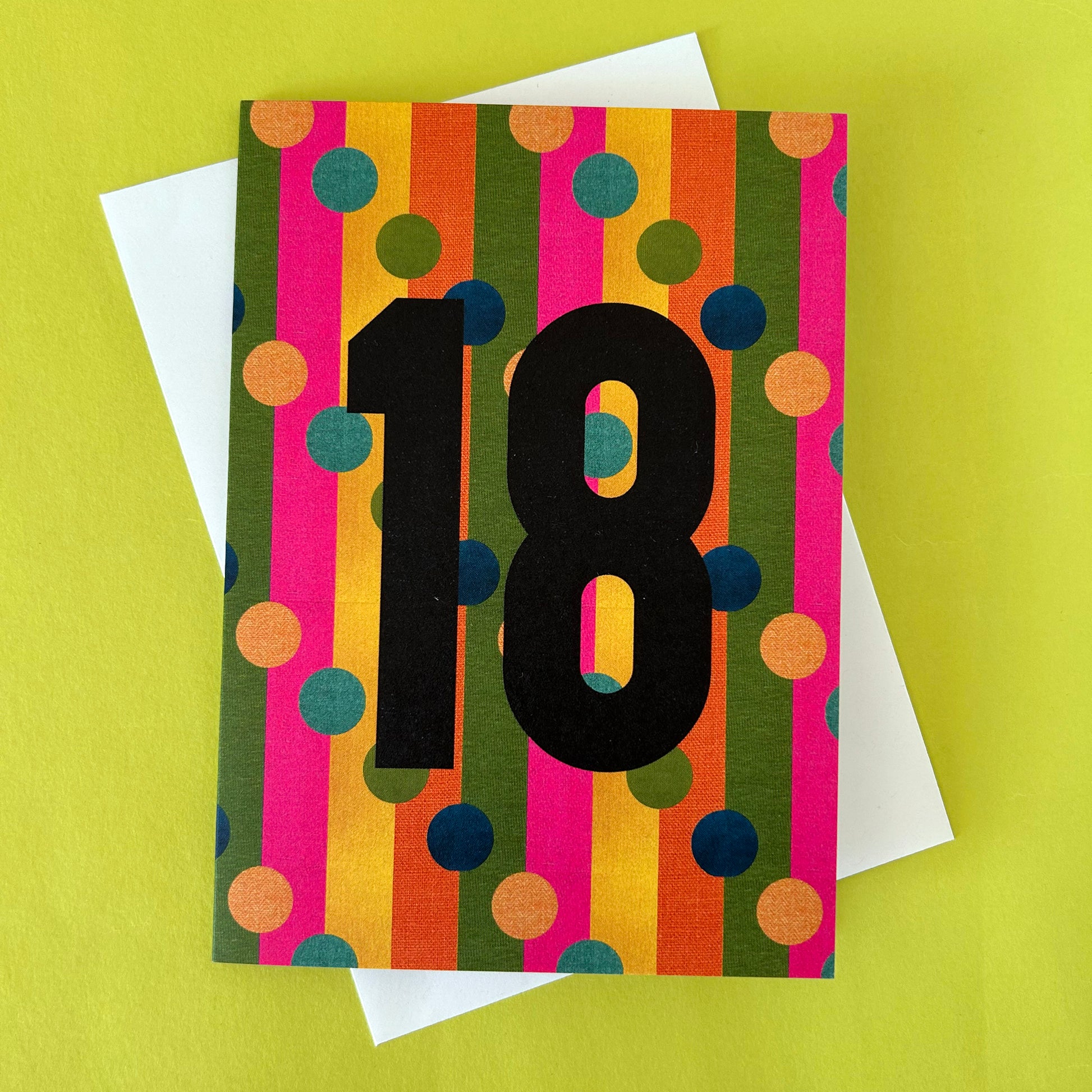 A colourful 18th birthday greeting card with bold black numerals '18' over vertical stripes of pink, orange, yellow, green, and blue polka dots, shown with a white envelope on a bright yellow-green background.